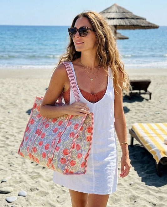 Woman on a beach holding a colorful tote bag, wearing a white dress.