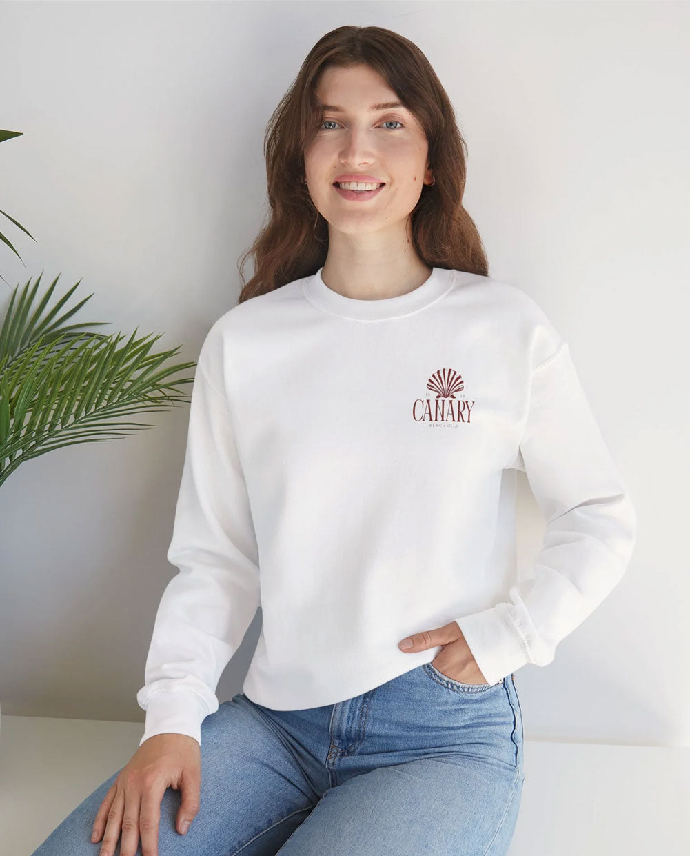 Woman wearing a white sweatshirt with a logo, sitting indoors with a plant in the background
